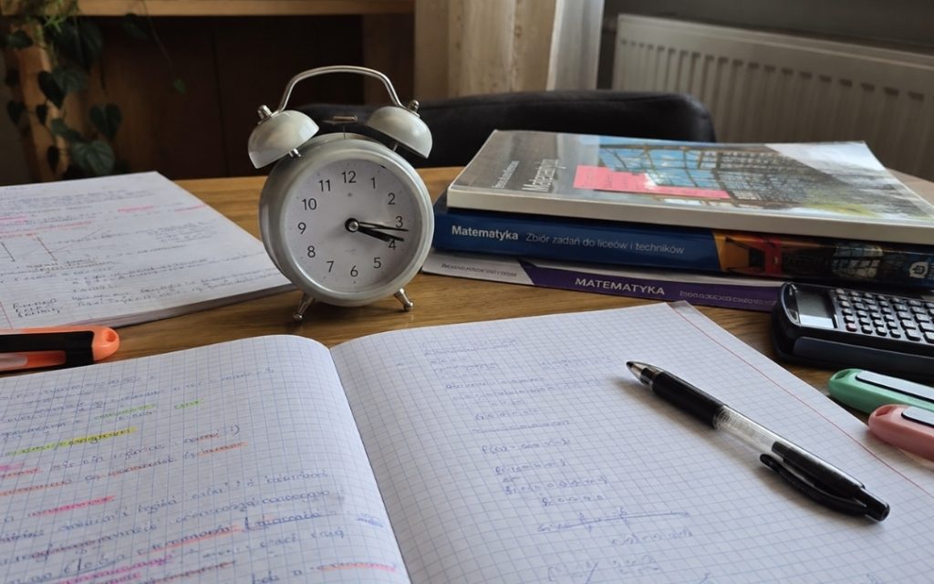 Budzik stoi na biurku obok otwartego zeszytu, długopisu, notatek i książek do matematyki.