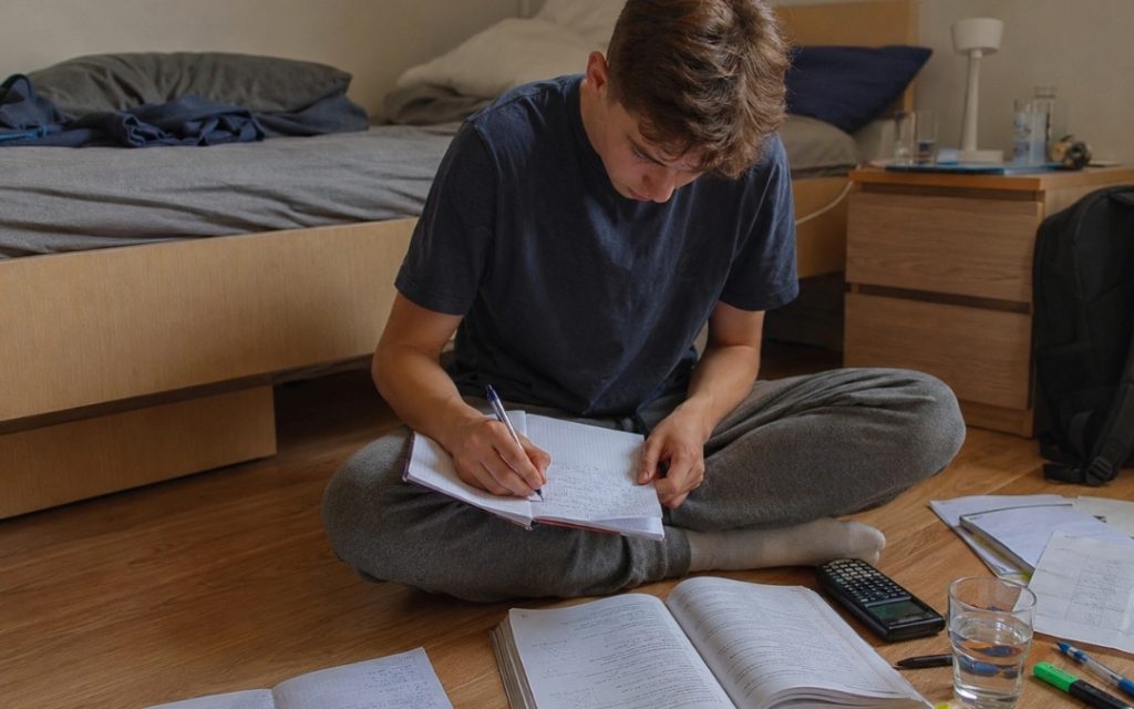 Chłopiec siedzi po turecku na podłodze w swoim pokoju, przed łóżkiem i uczy się matematyki.