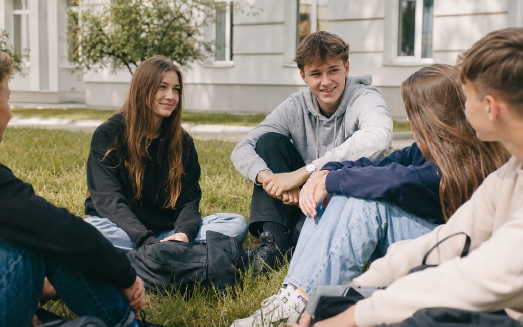 Grupa uczniów siedzi na trawie przed szkołą podczas przerwy.