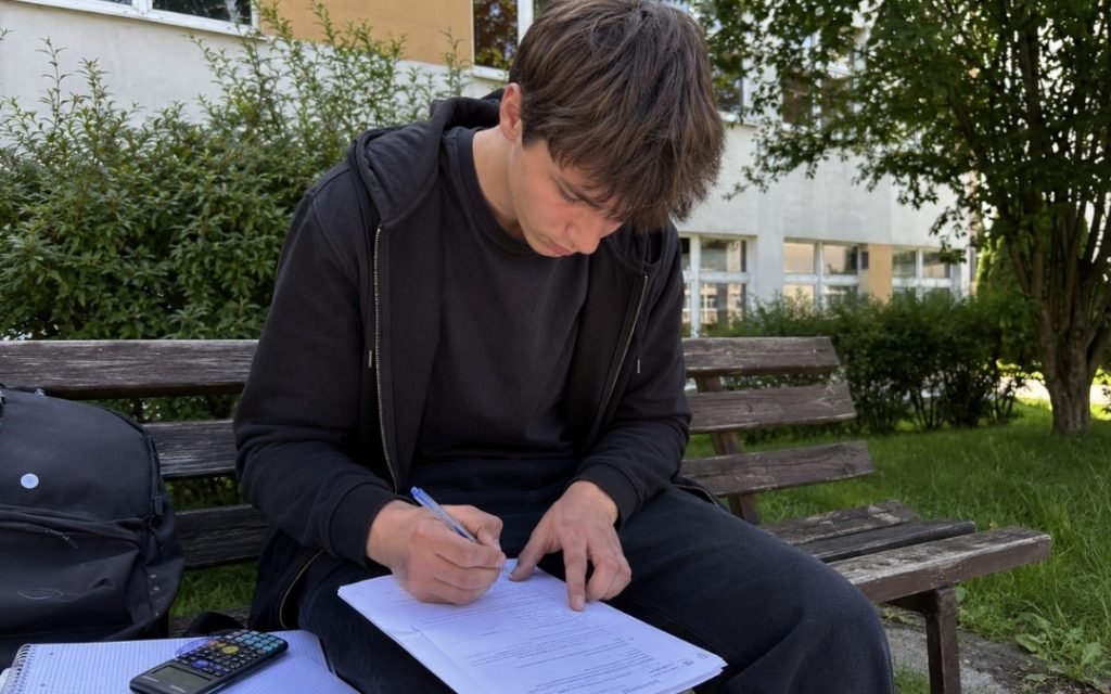 Maturzysta siedzi na ławce przed szkołą wśród zieleni i rozwiązuje kartę pracy z matematyki.