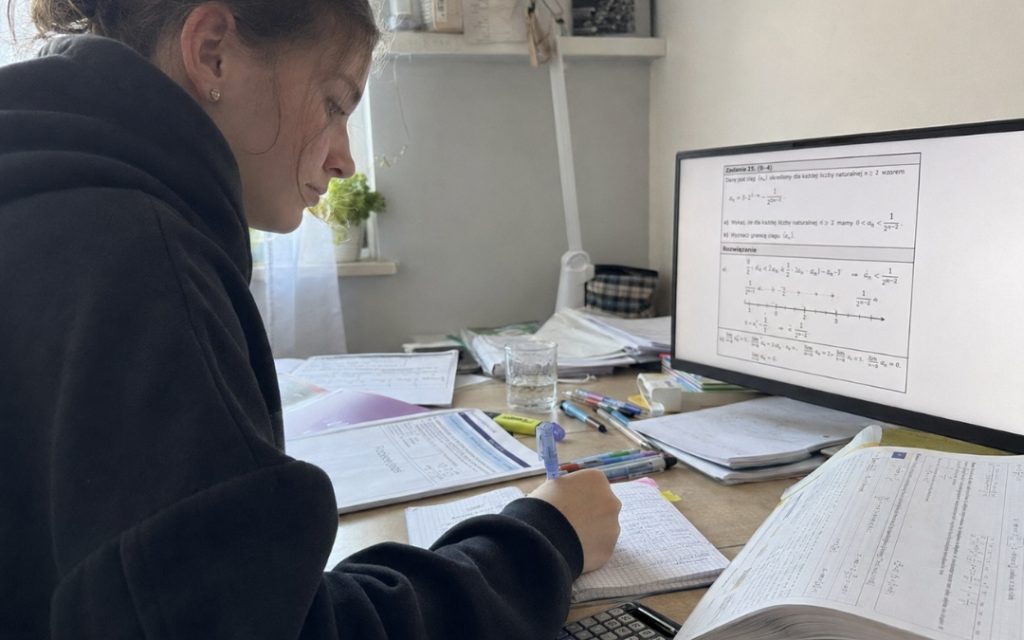 Nastolatka uczy się matematyki przy biurku, notując w zeszycie obok komputera z rozwiązaniem zadania maturalnego.
