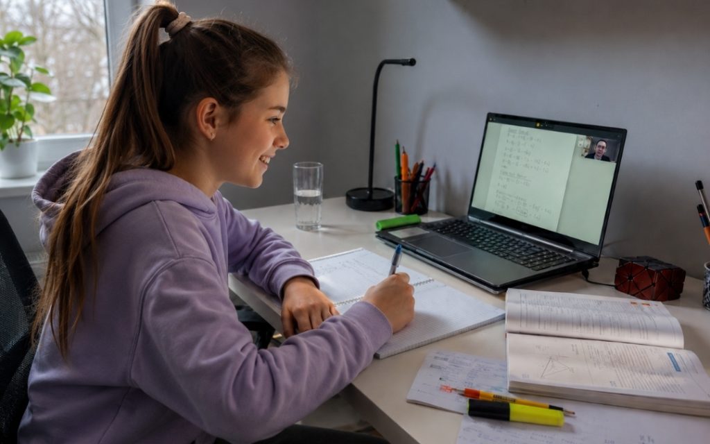 Uśmiechnięta uczennica podstawówki uczy się online przy laptopie i notuje w zeszycie.