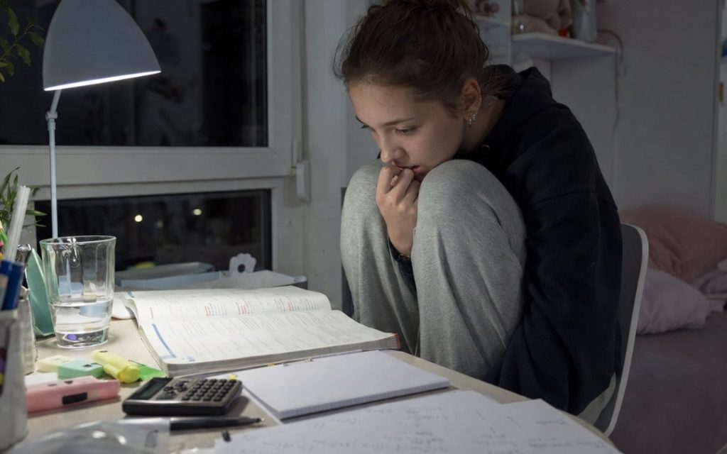 Zestresowana nastolatka siedzi przy biurku z kolanami pod brodą i patrzy na otwarty podręcznik do matematyki.