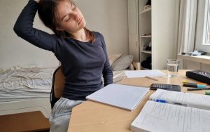 Nastolatka robi przerwę od nauki i rozciąga szyję przy biurku z zeszytem, podręcznikiem i kalkulatorem.