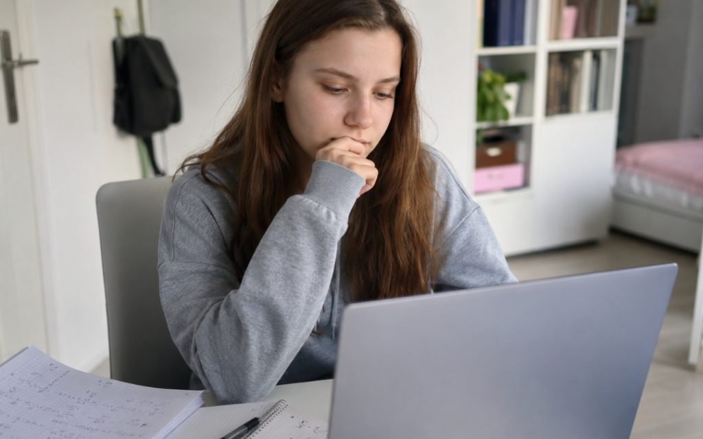 Nastolatka siedzi przed laptopem i analizuje notatki z matematyki, trzymając dłoń przy twarzy w skupieniu.