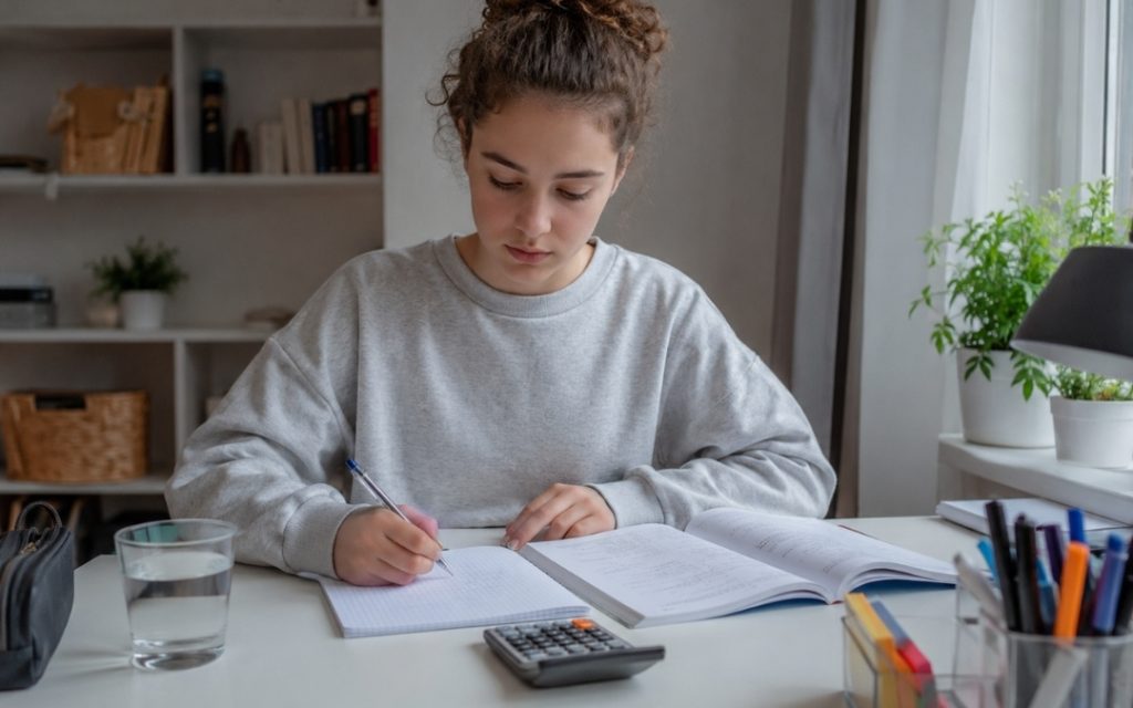 Nastolatka spokojnie uczy się matematyki przy biurku, zapisując notatki w zeszycie obok podręcznika, kalkulatora i szklanki wody.