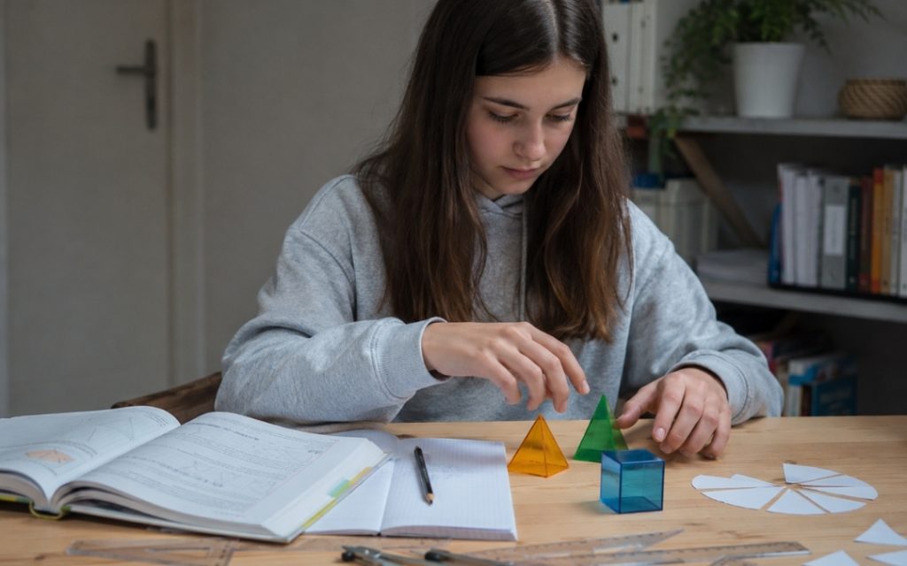 Nastolatka uczy się geometrii, układając bryły i pomoce matematyczne przy stole z zeszytem oraz podręcznikiem.