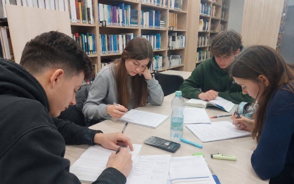 Licealiści uczą się razem w bibliotece, zapisując notatki i rozwiązując zadania przy wspólnym stole.