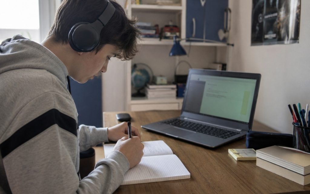 Nastolatek uczy się online przy laptopie w słuchawkach, zapisując notatki w zeszycie.