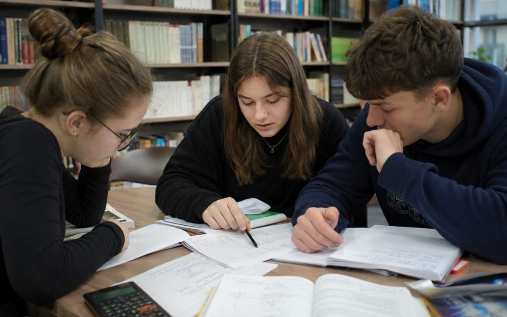 Trójka licealistów uczy się razem w bibliotece, omawiając zadania i zapisując rozwiązania w zeszytach.