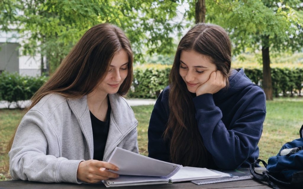 Dwie licealistki uczą się razem w plenerze, przeglądając notatki przy drewnianym stole w parku.