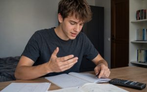 Licealista analizuje materiał z matematyki przy biurku, gestykulując i zapisując własne objaśnienia w zeszycie.