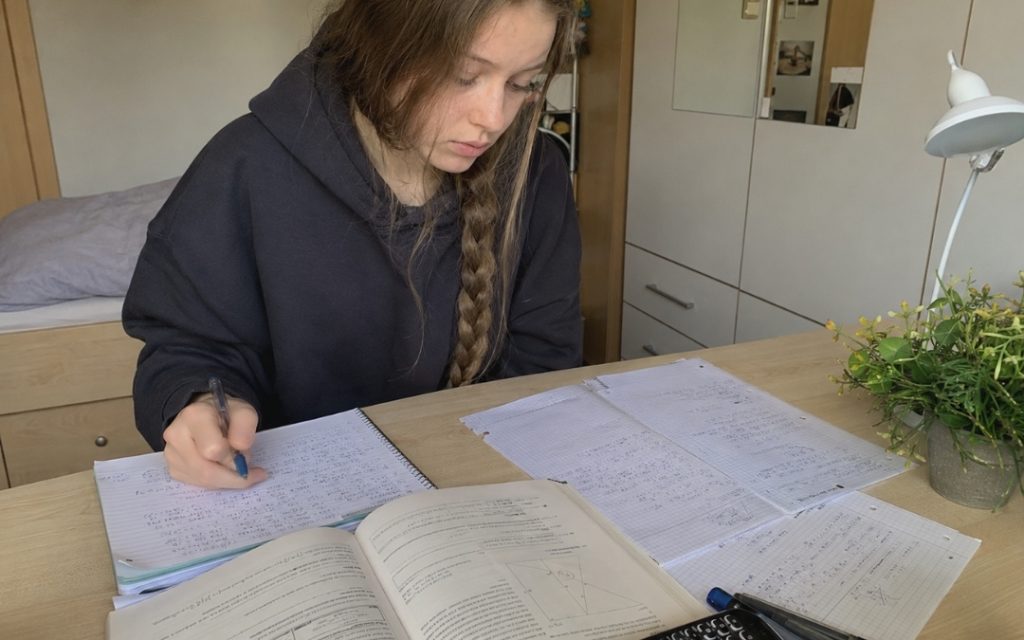 Licealistka uczy się matematyki przy biurku, przepisując i upraszczając notatki na kartkach oraz w zeszycie.
