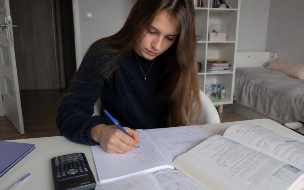 Licealistka uczy się matematyki przy biurku, zapisując notatki w zeszycie obok podręcznika i kalkulatora.