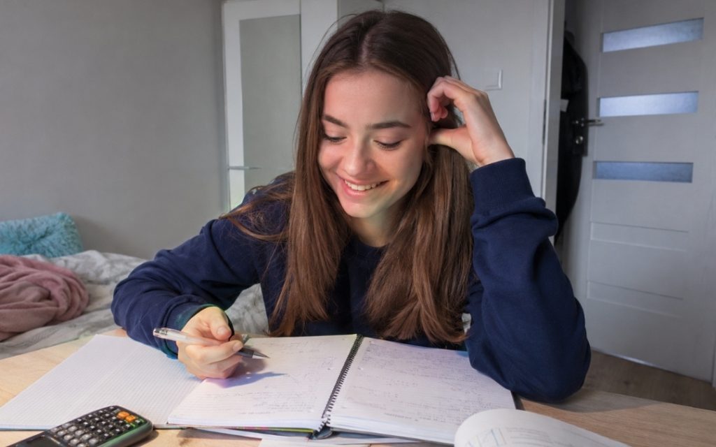 Uśmiechnięta licealistka siedzi przy biurku i z satysfakcją przegląda własne notatki z matematyki po zrozumieniu materiału.