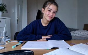 Uśmiechnięta licealistka siedzi przy biurku z gotowymi notatkami metodą Cornella, podręcznikami i przyborami do nauki.