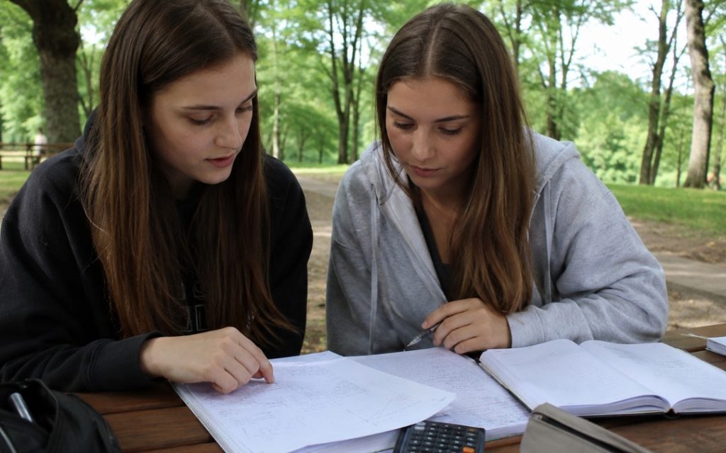 Dwie licealistki uczą się matematyki w plenerze, omawiając notatki i zadania przy drewnianym stole.
