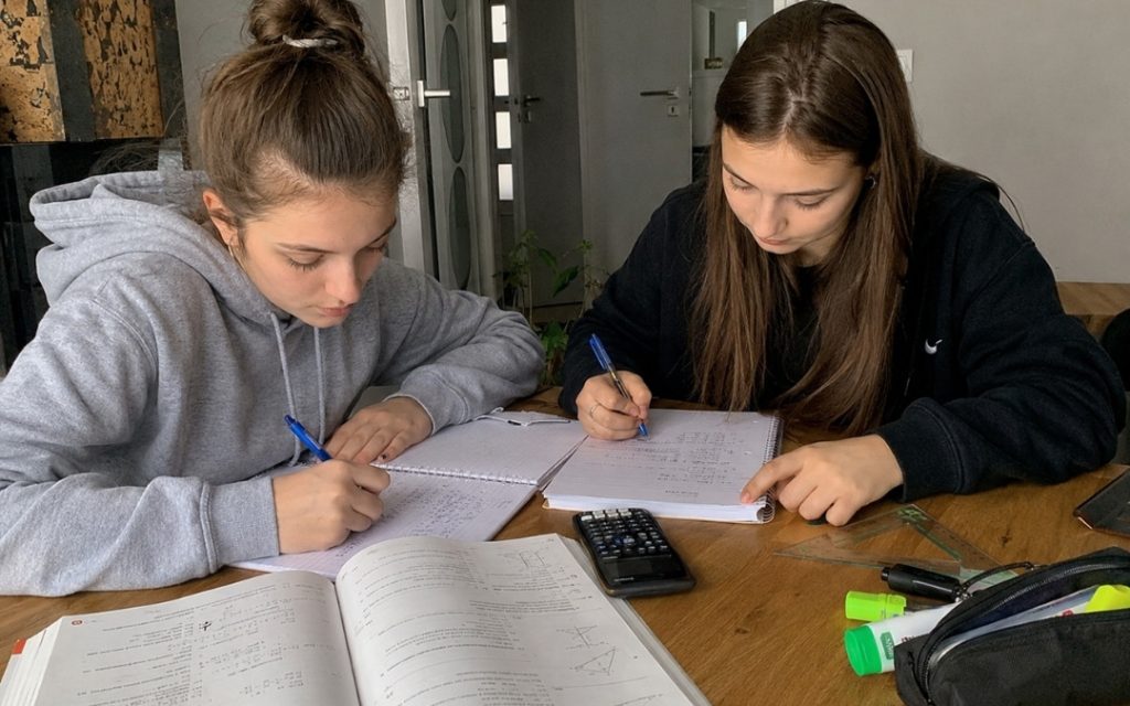 Dwie licealistki wspólnie rozwiązują zadania matematyczne przy stole, zapisując obliczenia w zeszytach.