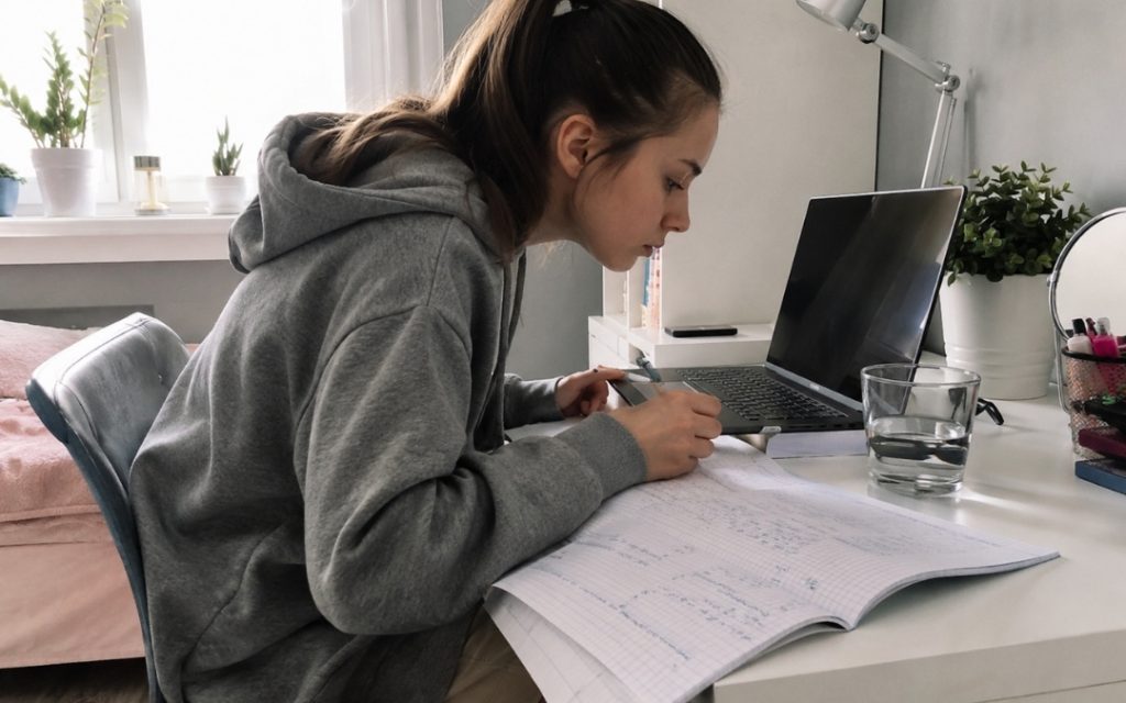 Nastolatka uczy się przy laptopie, zapisując notatki w zeszycie na biurku z podręcznikami i szklanką wody.