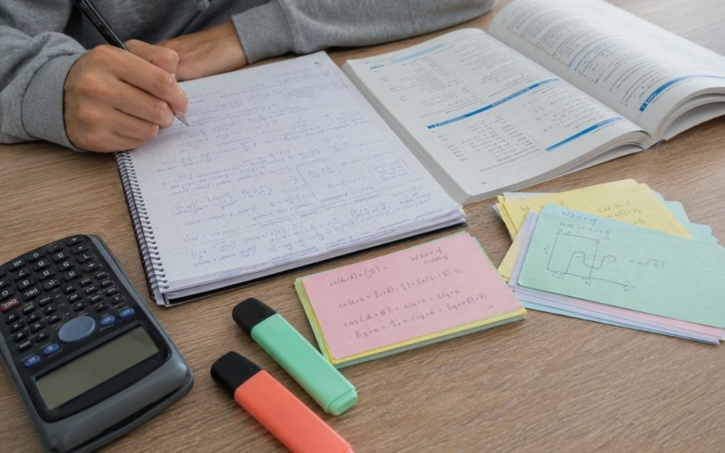 Zbliżenie na biurko z zeszytem, fiszkami, podręcznikiem, kalkulatorem i zakreślaczami podczas nauki matematyki.