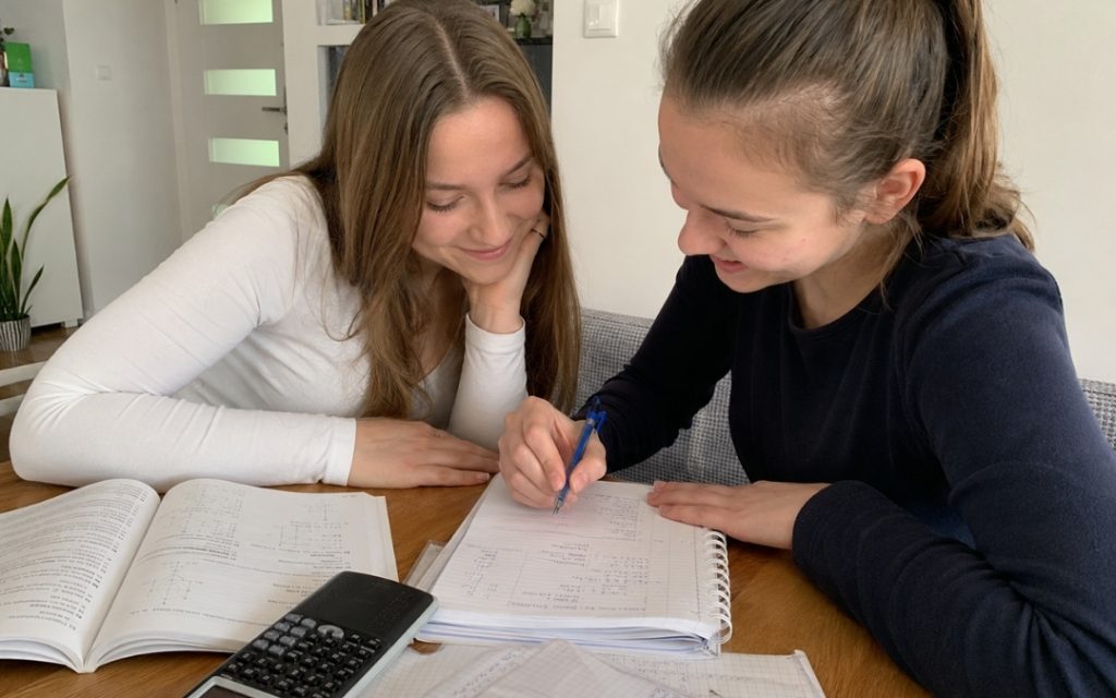 Dwie licealistki uczą się razem matematyki przy stole, uśmiechając się podczas omawiania zadania i notatek.