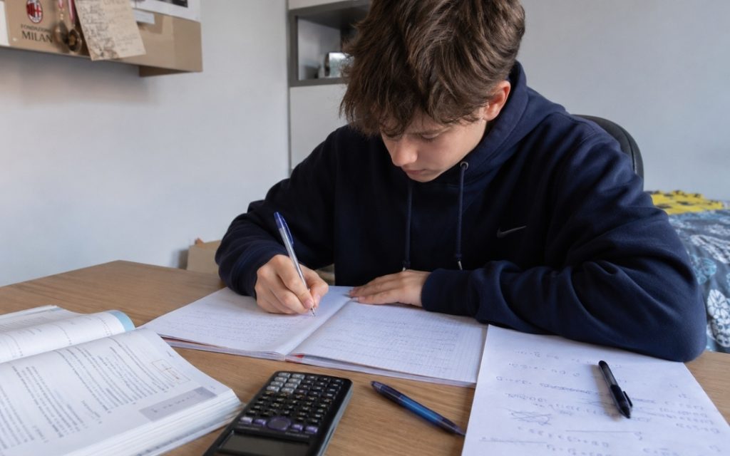 Licealista rozwiązuje zadania matematyczne przy biurku, korzystając z zeszytu, podręcznika i kalkulatora.