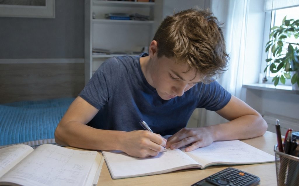 Licealista skupiony nad zadaniem matematycznym zapisuje rozwiązanie w zeszycie przy biurku z podręcznikiem i kalkulatorem.