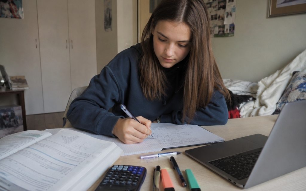 Licealistka pewnie uczy się matematyki przy biurku, zapisując obliczenia w zeszycie obok laptopa, podręcznika, kalkulatora i zakreślaczy.