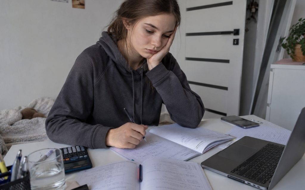 Licealistka siedzi przy biurku nad zeszytami i podręcznikiem do matematyki, wygląda na zablokowaną i zmęczoną nauką.