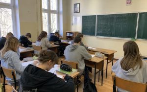 Uczniowie siedzą w ławkach w klasie i piszą w zeszytach, a na zielonej tablicy widać zapisane równania matematyczne.