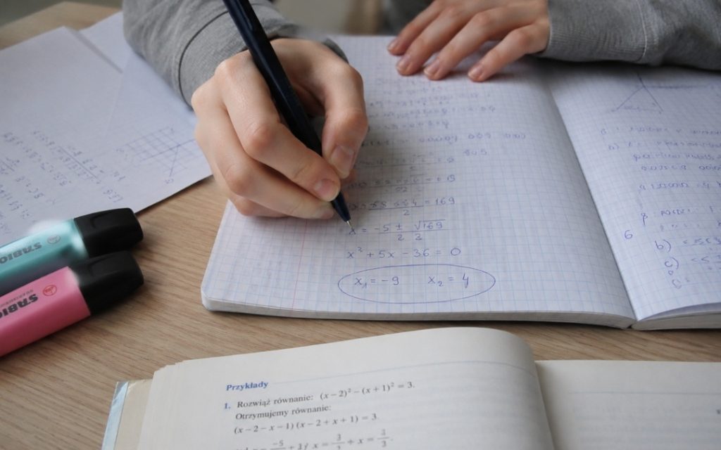 Dłonie nastolatki zapisują wzory matematyczne w zeszycie i zaznaczają wynik w kółko.