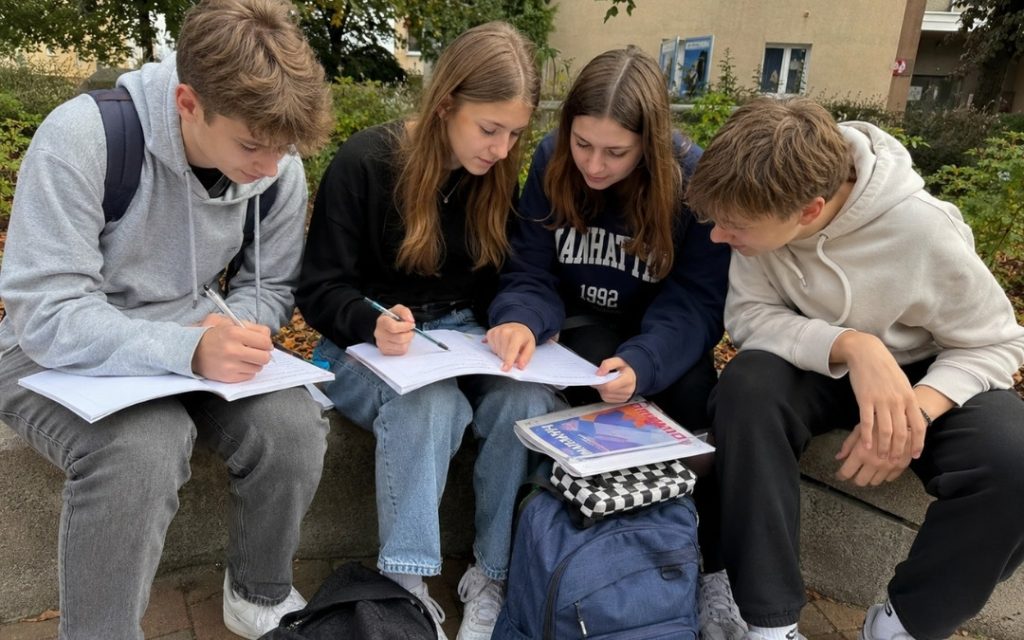 Czworo nastolatków siedzi na murku przed szkołą i wspólnie notuje w zeszytach.