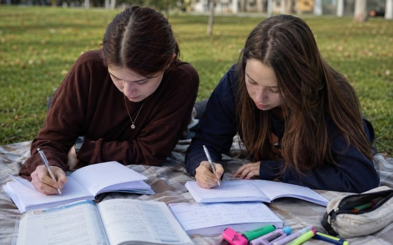 Dwie nastolatki leżą na brzuchu na kocyku na trawie i zapisują notatki w zeszytach.