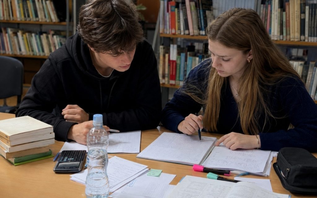 Dwójka nastolatków przygotowuje się do egzaminu w szkolnej bibliotece, korzystając z zeszytów, notatek i kalkulatora.