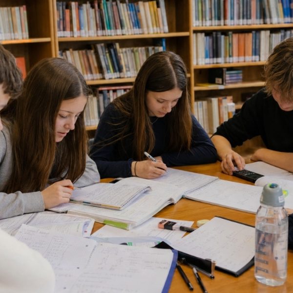 Grupka nastolatków uczy się razem przy stole w szkolnej bibliotece, korzystając z zeszytów, notatek i kalkulatora.