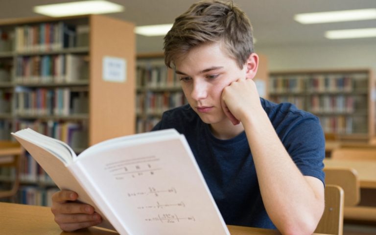 Nastolatek siedzi sam w szkolnej bibliotece i czyta gruby podręcznik do matematyki.