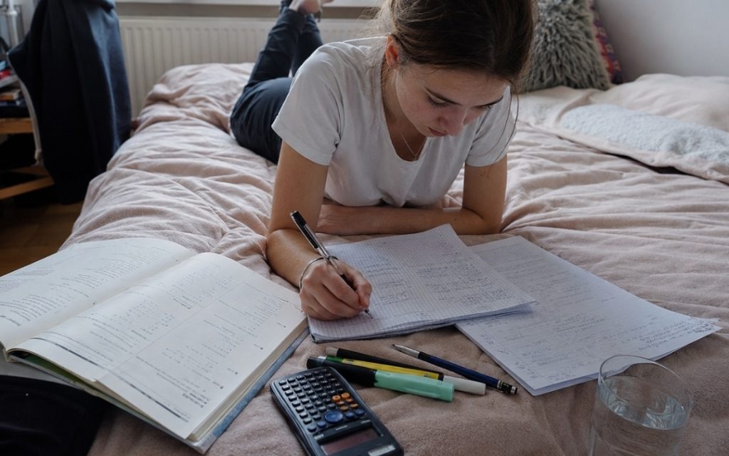 Nastolatka leży na brzuchu na łóżku i uczy się matematyki, notując w zeszycie obok podręcznika i kalkulatora.