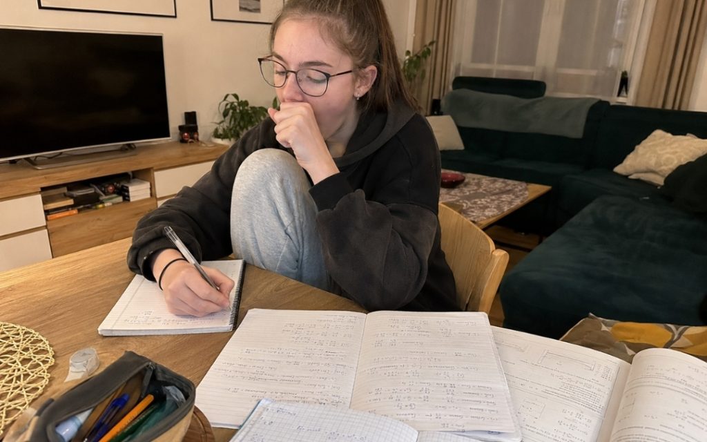Nastolatka w okularach i czarnej bluzie siedzi przy stole w salonie i notuje w zeszycie, otoczona notatkami z matematyki.