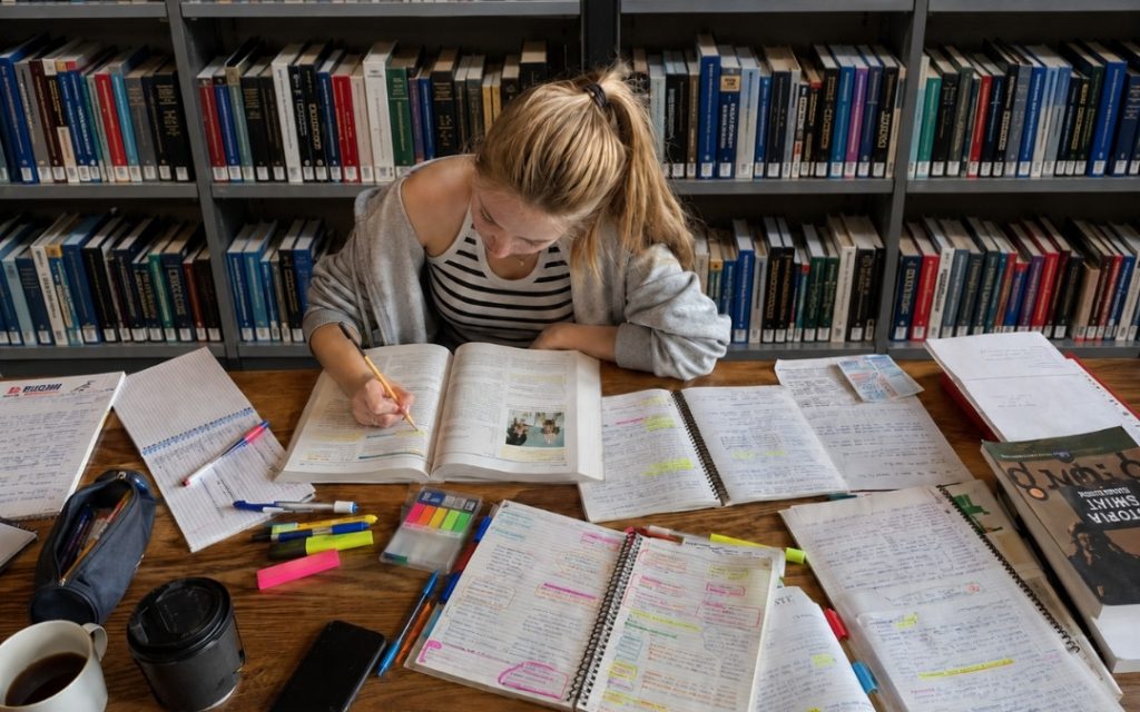 Nastolatka w pasiastej bluzce pochyla się nad otwartymi książkami i notatkami w bibliotece, otoczona zakreślaczami i kawą.