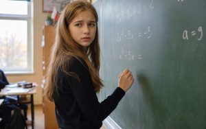 Nastolatka z niepewną miną stoi przy zielonej tablicy i pisze kredą równania matematyczne.