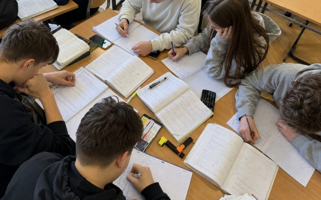 Nastolatkowie w klasie notują zadania w zeszytach, siedząc razem przy stołach widzianych z góry.