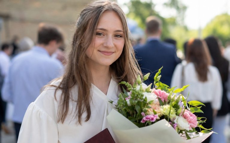 Szczęśliwa nastolatka w eleganckim stroju trzyma bukiet kwiatów podczas uroczystości szkolnej.