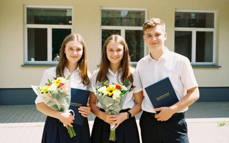 Troje absolwentów w strojach galowych stoi przed szkołą z bukietami kwiatów i granatowymi teczkami ze świadectwami.