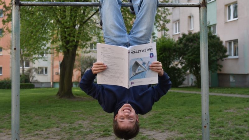 Uśmiechnięty chłopiec wisi do góry nogami na drążku i czyta podręcznik do matematyki.