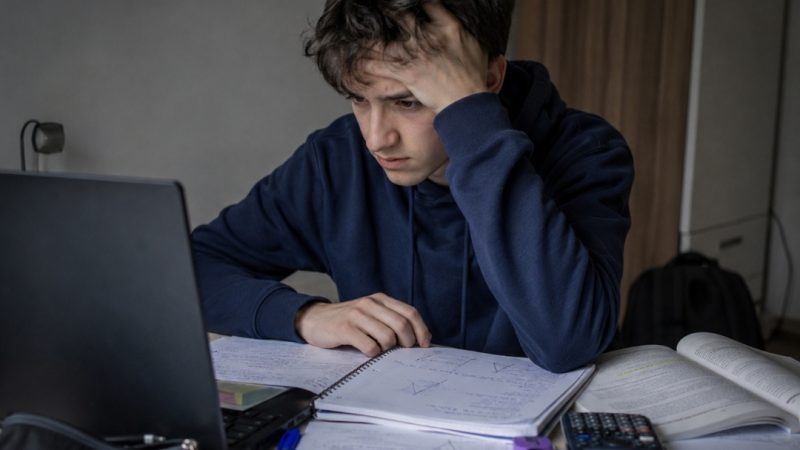Zestresowany maturzysta siedzi przed laptopem i patrzy na ekran, otoczony notatkami, zeszytami i materiałami do nauki.