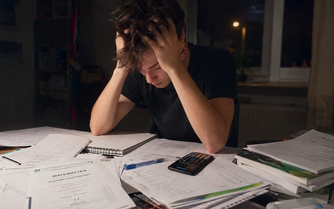 Zestresowany nastolatek łapie się za głowę wieczorem przy biurku zawalonym arkuszami z matematyki, kalkulatorem i podręcznikami.