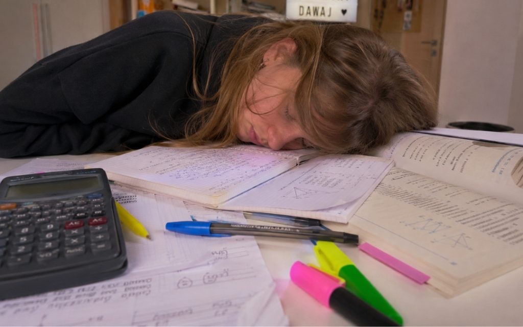 Zmęczona nastolatka zasnęła z głową na otwartym zeszycie z notatkami z matematyki, otoczona kalkulatorem, zakreślaczami i długopisami na biurku.