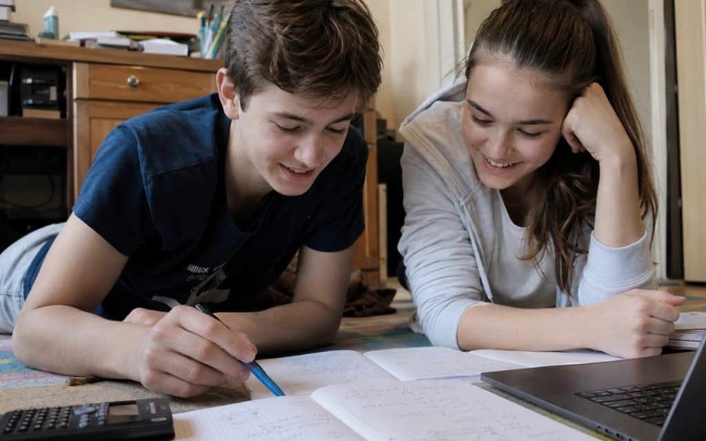 Dwoje licealistów uczy się razem matematyki na podłodze w pokoju, korzystając z laptopa i notatek.
