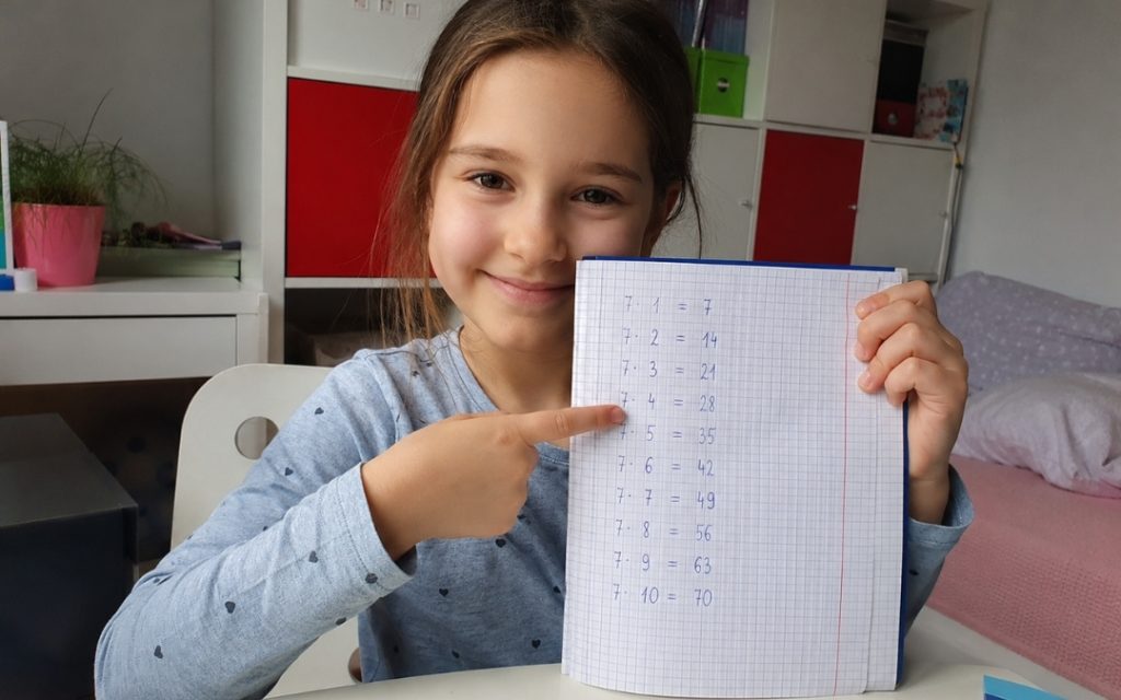 Uśmiechnięte dziecko dumne z rozwiązanych zadań matematycznych siedzi przy biurku w domu.