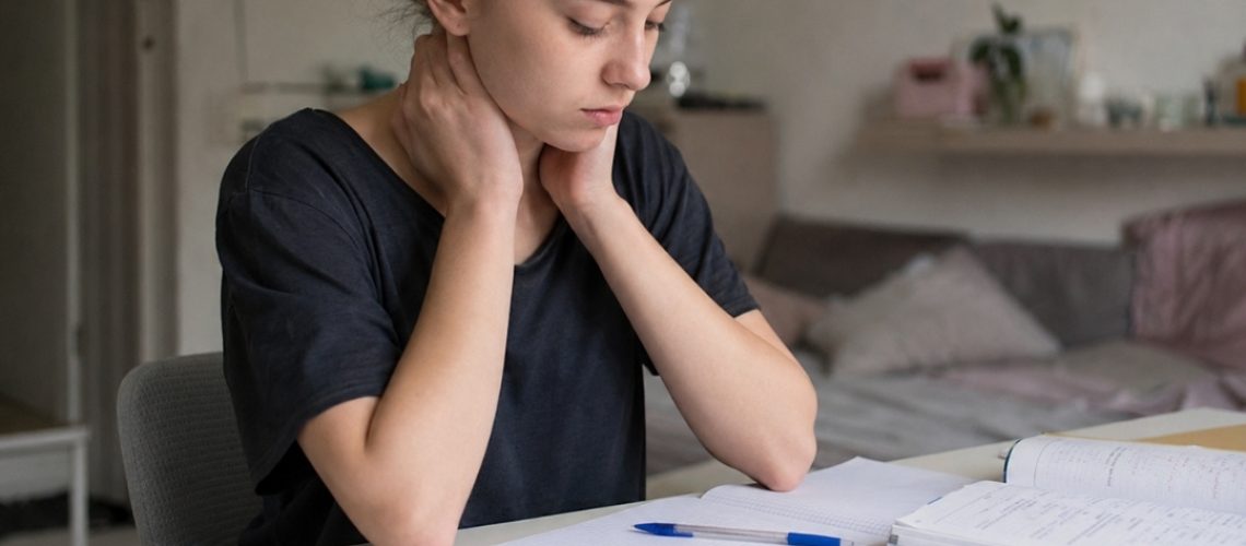 Licealistka siedzi przy biurku nad notatkami z matematyki i trzyma dłonie na karku, pokazując zmęczenie po długiej nauce.